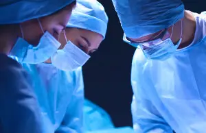 Two surgeons in blue scrubs and surgical masks leaning in with focused concentration during an operative procedure against a dark background