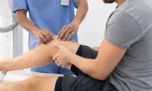 Clinician in blue scrubs performing a hands-on knee examination on a male patient seated on an exam table in a radiology or imaging suite
