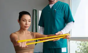Physical therapist in teal scrubs guiding a female patient through resistance band exercises during a post-surgical rehabilitation session